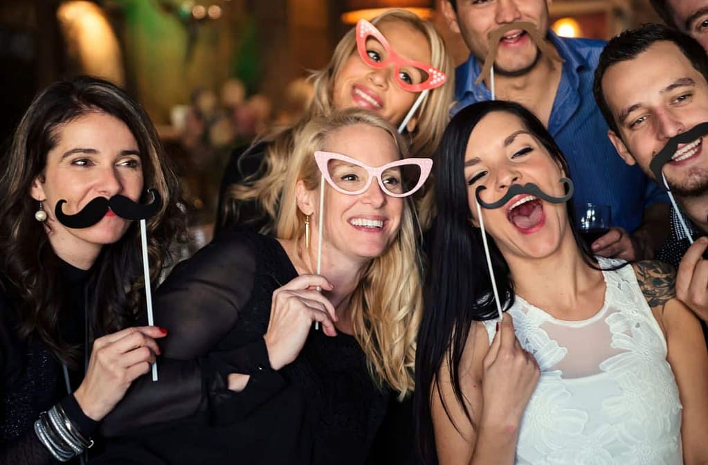 Group of friends laughing and posing with mustache and glasses props in a photo booth.