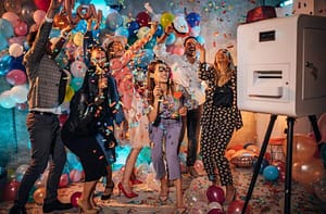Group of people celebrating with props, confetti by a photo booth surrounded by balloons.