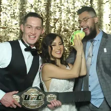 Three friends posing in a photo booth with a UFC belt prop and a playful taco prop against a gold backdrop.