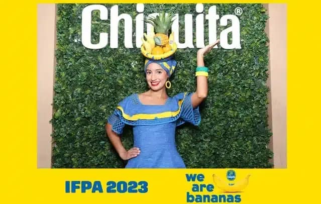 Smiling lady in a fruit-themed headpiece posing in a photo booth brand activation with an artificial green leaves backdrop.