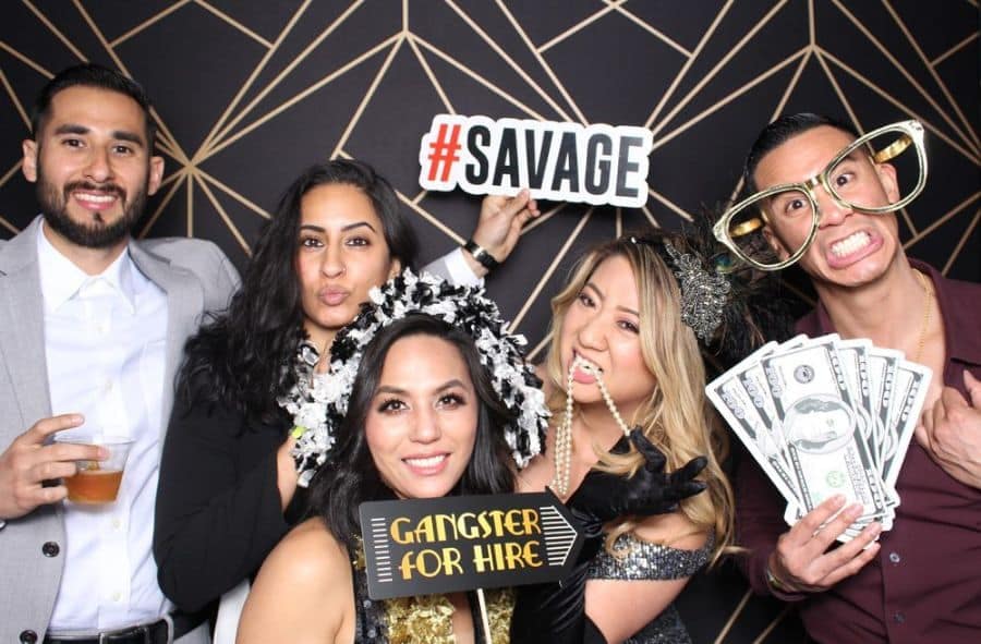 Group of friends posing with playful props like #SAVAGE sign, money fan, and Gangster for Hire in front of geometric backdrop.