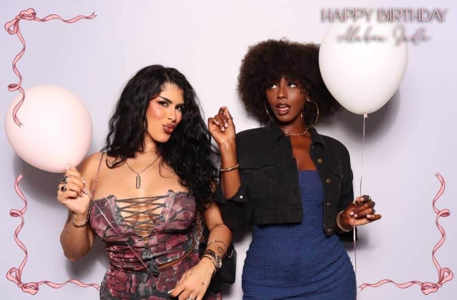 Two women posing in a birthday photo booth with balloons and playful expressions.